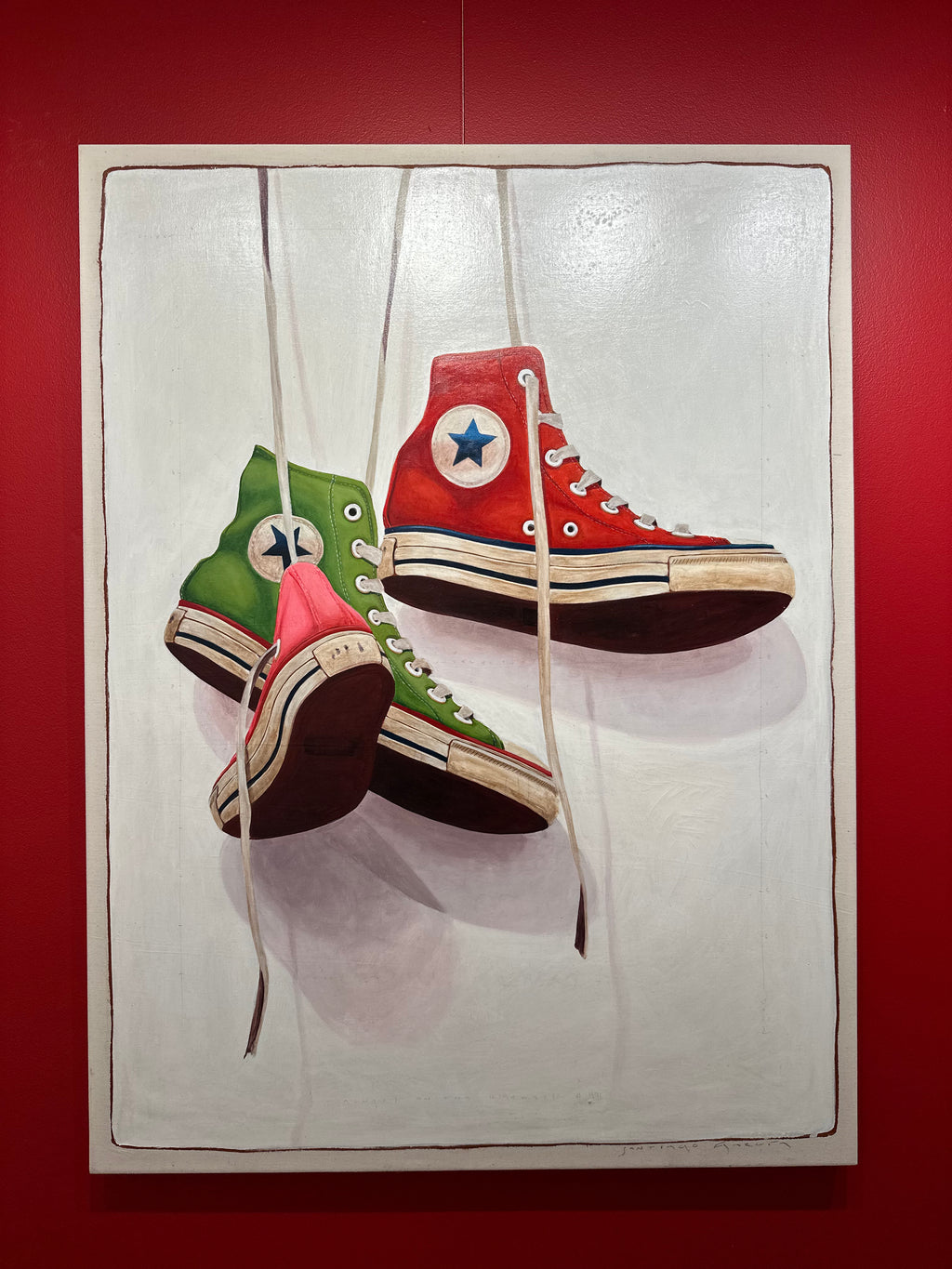 Still life oil painting by Santiago Garcia of a red, green, and pink converse sneaker hanging by shoe laces on a white shadowed canvas