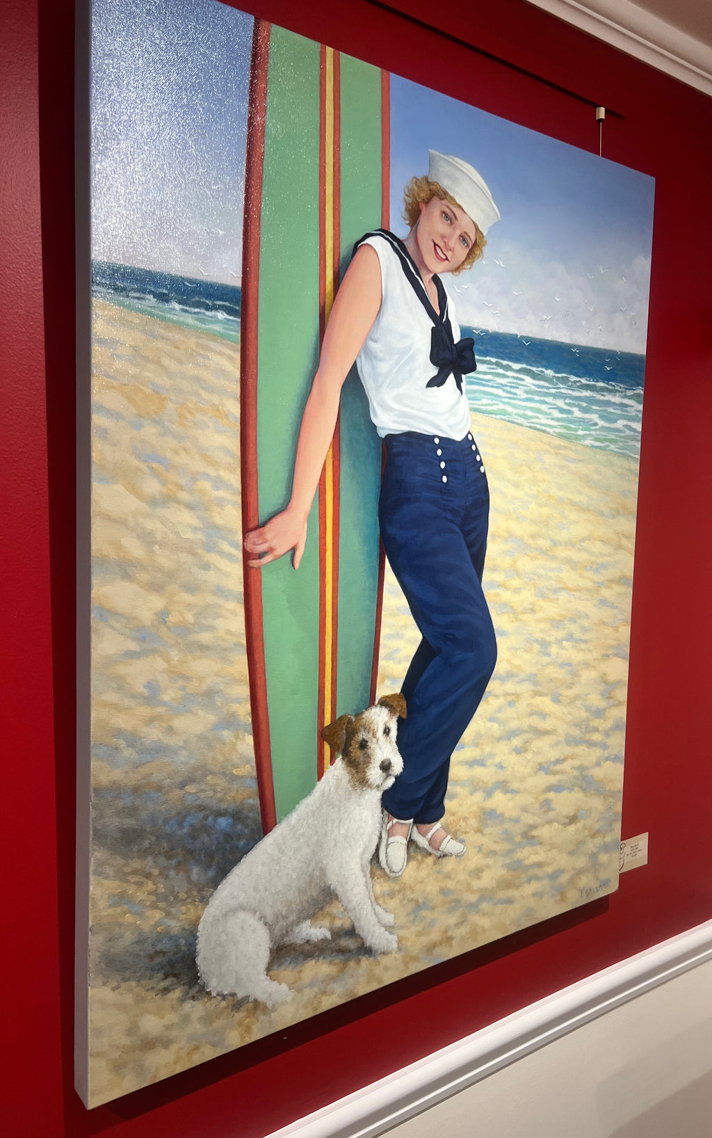 oil painting of a blonde woman with navy sailor pants, top and hat staning in front of a surfboard on the beach with terrier dog