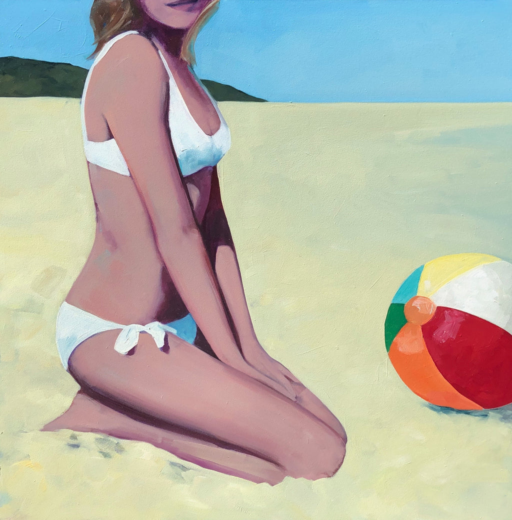 Blonde woman in a white bikini kneeling on the sand with a beach ball in front of her. Green landscape and blue sky background.